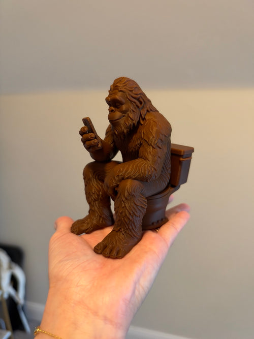 Brown 3D printed Bigfoot figurine sitting on a toilet held in hand